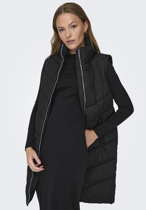 Veste rembourrée noire avec un col haut, dotée d'une fermeture éclair et d'un motif matelassé, portée sur une robe noire à manches longues.