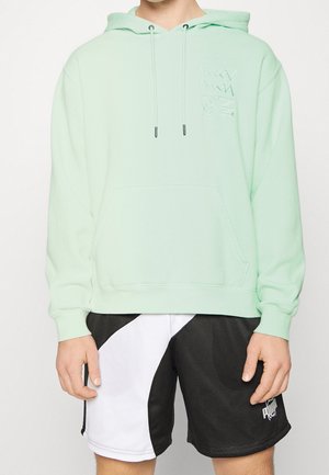 Mężczyzna w miętowo-zielonej bluzie z kapturem z subtelnym logo Puma oraz czarno-białych spodenkach do koszykówki Puma na jednolitym białym tle.