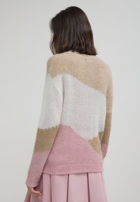 Donna con capelli scuri lunghezza spalle che indossa un maglione a blocchi di colore beige, bianco e rosa e una gonna plissé rosa chiaro, girata di spalle.