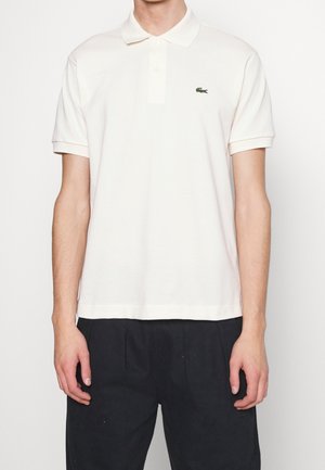 Mann trägt ein schlichtes weißes kurzärmeliges Poloshirt mit einem kleinen grünen Krokodil-Logo auf der linken Brustseite und schwarze Hose.