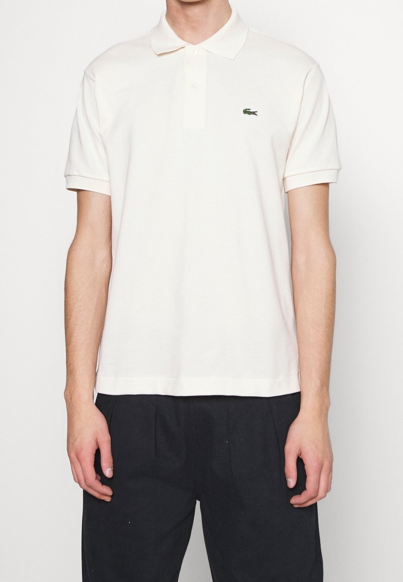 Homme portant un polo blanc uni à manches courtes avec un petit logo crocodile vert sur la poitrine gauche et un pantalon noir.