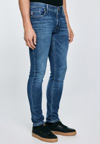 Férfi, aki keskeny szabású kék farmert és fekete tornacipőt visel, barna talppal, világos, egyszínű háttér előtt áll.