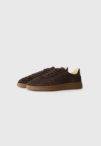 Brown suede sneakers with a textured upper, round toe, and tonal laces. Features a rubber sole and subtle branding on the side.