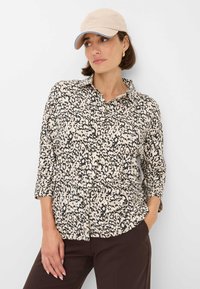 Camicia a maniche lunghe con stampa leopardata in crema e nero, dotata di colletto e maniche a tre quarti. Indossata con pantaloni marroni e un cappellino beige.
