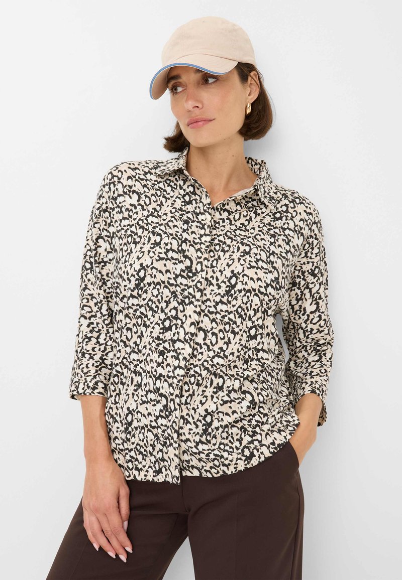 Camicia a maniche lunghe con stampa leopardata in crema e nero, dotata di colletto e maniche a tre quarti. Indossata con pantaloni marroni e un cappellino beige.