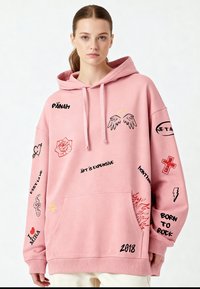 Hoodie surdimensionné rose en matériau doux, avec divers graphismes et textes en noir et rouge, y compris des ailes, des cœurs et une croix.