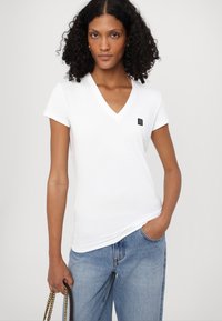 Camiseta blanca de cuello en V hecha de algodón, con mangas cortas, que presenta un pequeño parche negro con logo en el lado izquierdo del pecho. La lleva puesta con jeans de color azul claro.