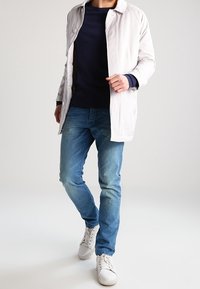 Světlá šedá bunda s knoflíkovým límcem, tmavě modrá košile a modré džíny. Bílé tenisky doplňují outfit, který má neformální design.