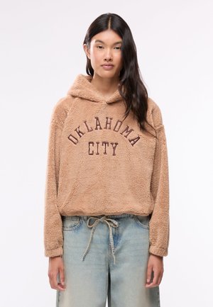 Giovane donna che indossa una felpa beige pelosa con testo "Oklahoma City" e jeans azzurri chiari annodati in vita, in piedi davanti a uno sfondo semplice.