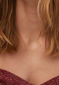 Vêtement en tricot bordeaux avec un décolleté en V, design ajusté et tissu texturé. Cheveux partiellement visibles, encadrant le décolleté.