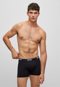 Schwarze Boxer-Shorts mit einem weißen Logo-Bund, aus glattem Stoff, enganliegendem Design und kurzen Beinen, getragen von einem Modell mit durchtrainiertem Körper.