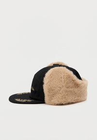 Svart caps med en tan fuskepelskant og bak. Har brodert gulldetaljering på fronten. Myk tekstur totalt sett, strukturert form.