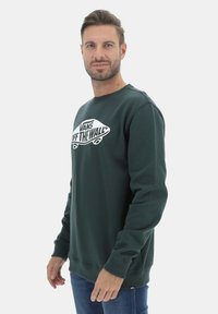 Felpa verde scuro con scollo a girocollo e polsini a costine. Presenta una grafica del logo bianco "Vans Off The Wall" sul fronte. Tessuto morbido.