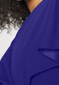 Un tejido azul real con una textura suave presenta acentos con volantes a lo largo del escote, destacando un diseño suave y fluido.