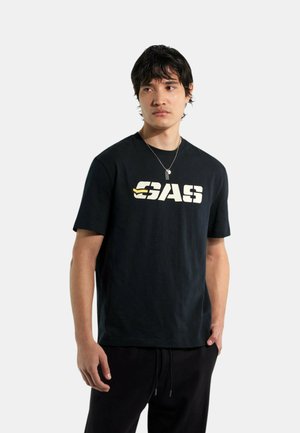 Giovane uomo che indossa una maglietta nera con il logo "GAS" in bianco e giallo, pantaloni neri e una collana d'argento, in piedi davanti a uno sfondo bianco.