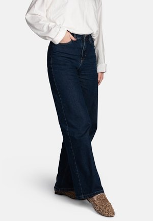 Person, die dunkelblaue, hochgeschnittene Jeans mit geradem Schnitt, ein weißes Langarmshirt und Loafer mit Leopardenmuster trägt und mit einer Hand in der Tasche steht.