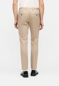 Άνδρας που φοράει μπεζ παντελόνι κοστουμιού στενής γραμμής, λευκές κάλτσες και μαύρα παπούτσια κοστουμιού, στέκεται γυρισμένος με την πλάτη σε λευκό φόντο.