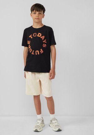 Zwarte katoenen t-shirt met cirkelvormige oranje tekst "DE TOEKOMST VANDAAG", gecombineerd met crèmekleurige shorts en grijze sportschoenen.