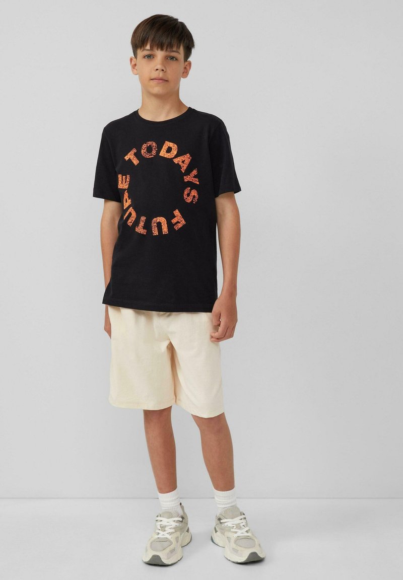 Schwarzes Baumwoll-T-Shirt mit kreisförmigem orangenem Schriftzug "DIE ZUKUNFT VON HEUTE", kombiniert mit cremefarbenen Shorts und grauen Sportschuhen.