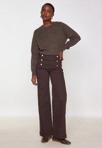 Femme portant un pantalon large marron foncé avec des boutons dorés, un pull marron assorti, des boucles d'oreilles dorées, et des chaussures marron, se tenant avec une main sur la hanche.