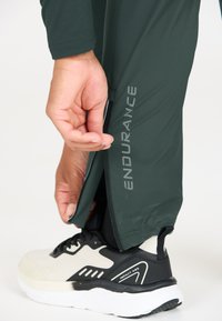 Groene sportbroek met zijrits, voorzien van reflecterende "ENDURANCE" tekst, gecombineerd met een zwart en crèmekleurige sneaker. Glad textuur van de stof.