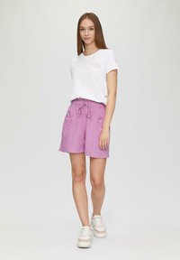 Witte T-shirt met een klein logo, gecombineerd met paarse shorts met een elastische tailleband en zijzakken. Model draagt witte sneakers.