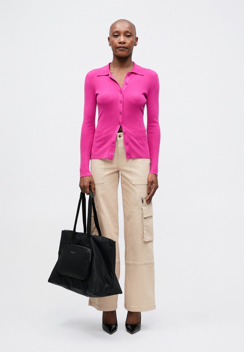 Cardigan rosa a coste con colletto, abbinato a pantaloni cargo beige. Borsa tote nera con una pochette più piccola. Il modello indossa decolté nere a punta.