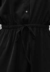 Κοντινό πλάνο ενός μαύρου φορέματος με ελαστική μέση που σφίγγει και δένει με φιόγκο, καθώς και μερικώς κουμπωμένο εμπρόσθιο μέρος με κουμπιά.