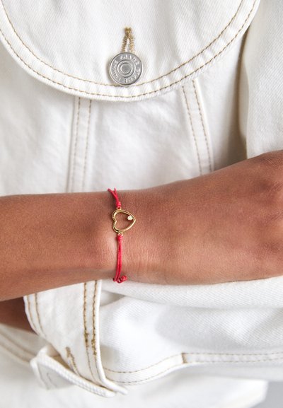 Sarkans auklas spilvens, kas rotāts ar zelta sirds formas šarmu un mazām apaļām akcentiem. Nēsāts uz plaukstas, pret gaišas krāsas auduma fonu.