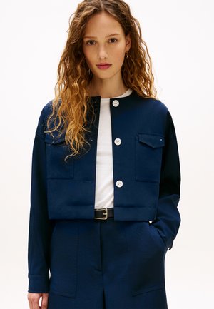 Frau mit lockigem Haar, die eine dunkelblaue, geknöpfte Jacke über einem weißen Hemd trägt, passende dunkelblaue Hose und einen schwarzen Gürtel, steht mit einer Hand in der Tasche.