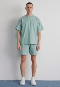 Tricou oversized din bumbac verde deschis și șorturi asortate. Design simplu, cu mâneci scurte și croială relaxată, purtat cu adidași albi.