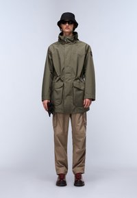 Olivgrüne wasserdichte Jacke mit Kapuze, verstellbarem Taillenbund und zwei vorderen Taschen, kombiniert mit beigefarbenen Cargo-Hosen und braunen Schnürstiefeln.