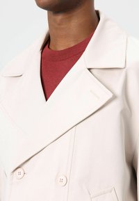 Beige dubbelgeknoopt jasje met grote ingesneden revers, twee knopen en schouderaccenten, gedragen over een bordeauxrood geribbelde trui.