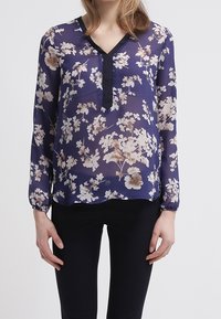 Blouse en voile navy avec un motif floral crème, décolleté en V et bord noir. Manches longues avec poignets élastiques, tissu léger. Pantalons noirs portés.