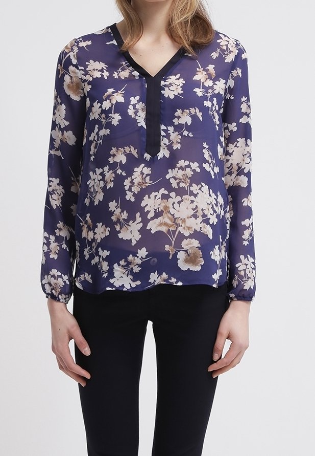 Blouse en voile navy avec un motif floral crème, décolleté en V et bord noir. Manches longues avec poignets élastiques, tissu léger. Pantalons noirs portés.