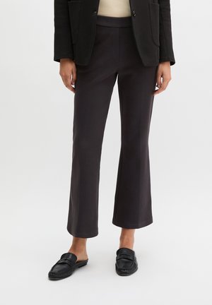 Vrouw draagt zwarte cropped wijde broek, zwarte loafers, beige top en zwarte blazer, staand tegen een effen achtergrond.