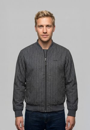 Lyshåret mand med kort hår iført en mørkegrå, stribet lynlåstrøje og jeans, stående mod en ensfarvet grå baggrund.