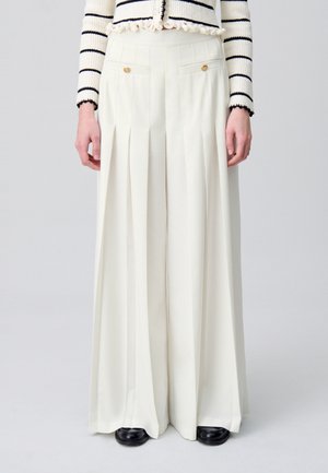 Pantalones plisados de pierna ancha en crema, con bolsillos laterales con acentos dorados. Tejido suave con forma estructurada; combinados con zapatos negros.