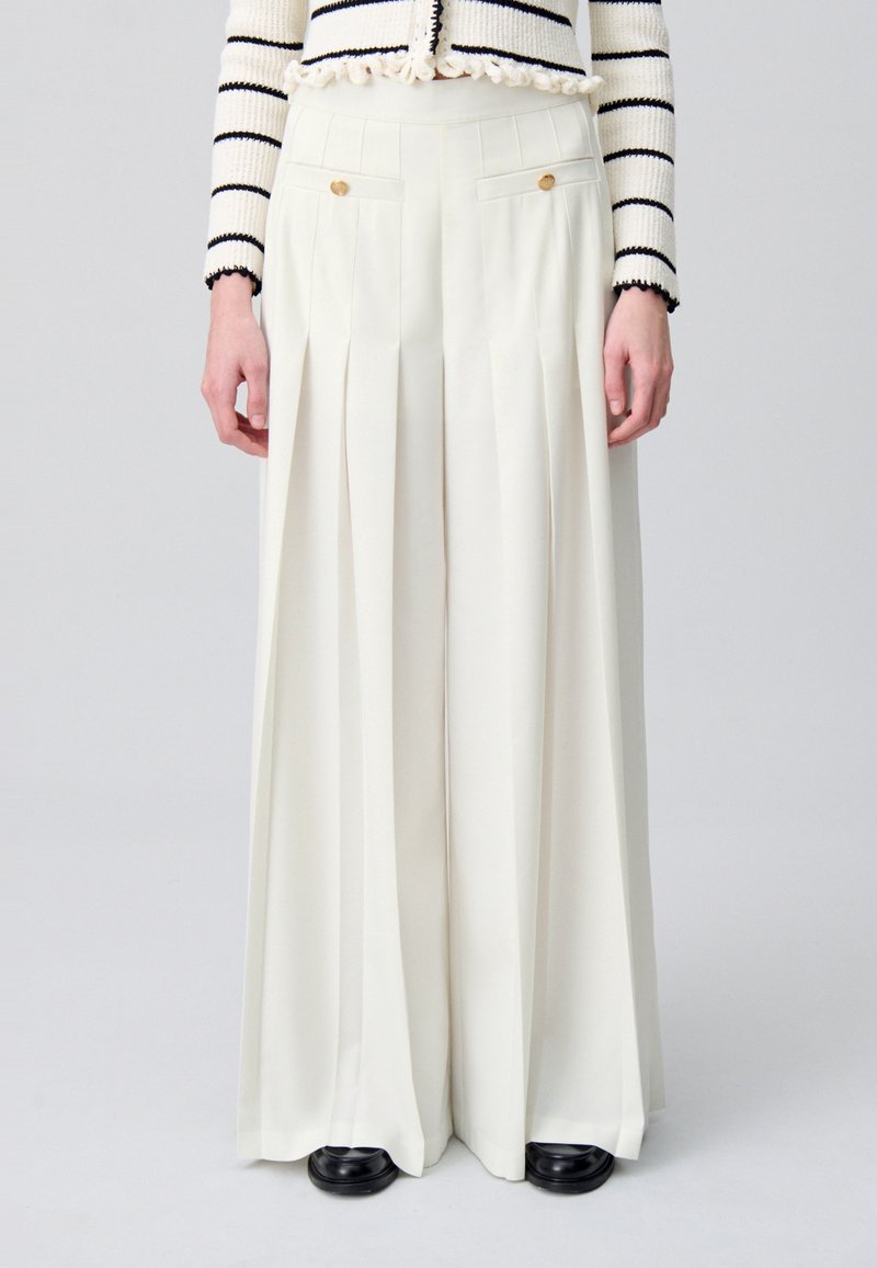 Pantalones plisados de pierna ancha en crema, con bolsillos laterales con acentos dorados. Tejido suave con forma estructurada; combinados con zapatos negros.