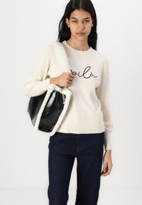 Pull crème avec "voilà" en script noir, poignets côtelés, accompagné d'un sac noir en verni avec un bord en fausse fourrure blanche et des bretelles. Jean sombre.