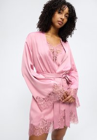 Γυναίκα με κατσαρά μαλλιά που φορά ρόμπα από ροζ σατέν με δαντέλα στα μανίκια, το τελείωμα και το ντεκολτέ, δεμένη στη μέση, στέκεται μπ ροστά σε ένα απλό φόντο.