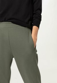 Olivgrüne Jogginghosen mit weicher Textur, seitlichen Taschen und einem elastischen Bund, kombiniert mit einem schwarzen Pullover mit gerippten Bündchen.