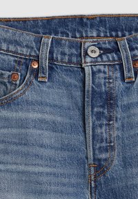 Vista frontal de la cinturilla de unos jeans de mezclilla azul que muestra el botón, los lazos para el cinturón, el bolsillo delantero y los remaches de cobre con costura naranja.