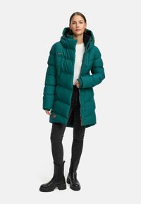 Turkusowa kurtka puchowa z wysokim kołnierzem, zamkiem błyskawicznym, bocznymi kieszeniami i guzikami jako akcentami. Połączona z czarnymi spodniami i masywnymi czarnymi botkami.