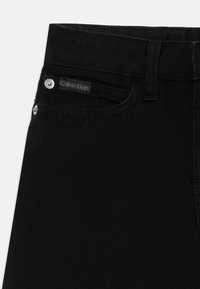 Schwarze Denim-Jeans mit einem Calvin Klein-Label auf der vorderen Tasche, silbernen Details und einem sauberen, strukturierten Finish.