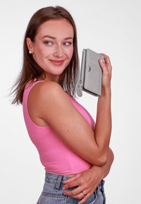 Šedá peněženka s texturovaným vzorem, se zipovým uzávěrem, držena proti růžovému tílku. Peněženka má hladké okraje a dekorativní táhlo zipu.