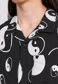 Chemise noire à manches courtes avec des motifs yin-yang blancs, un col à crans, une petite poche et une fermeture à boutons sur le devant. Texture de tissu lisse.