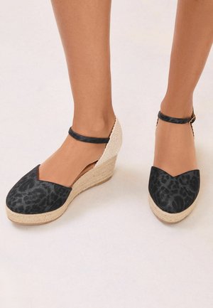 Wedge espadrille sandals with black leopard print closed toes, ankle straps, and woven beige platform heels on bare feet.