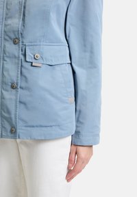 Hellblaue Jacke mit glatter Textur, ausgestattet mit Druckknöpfen und einer aufgenähten Tasche auf der linken Seite. Lange Ärmel und gerade geschnitten.