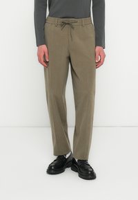 Person iført olivenfarvede snorebukser, sorte loafers, hvide sokker og en mørk langærmet bluse, der står mod en hvid baggrund.